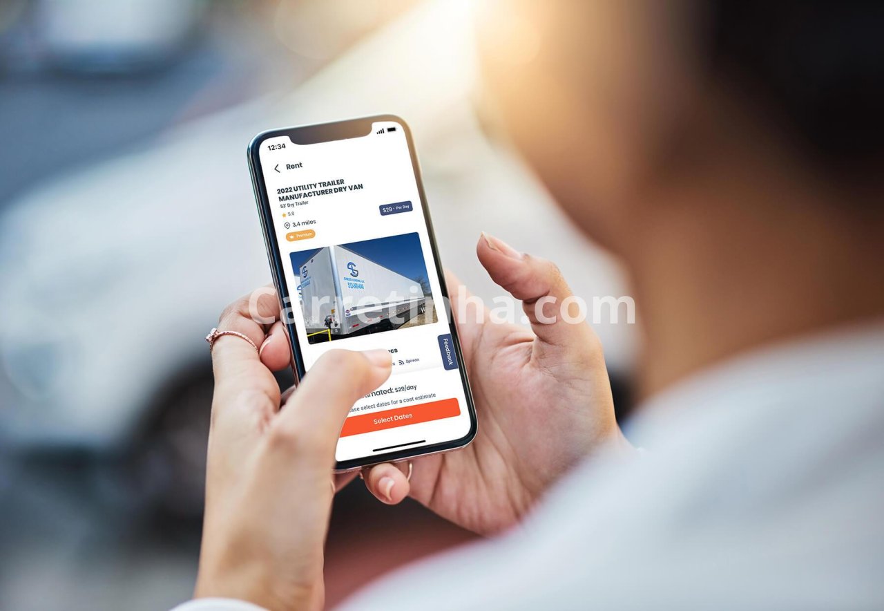Consumidor utiliza o smartphone para consultar opções de locação ao lado de um reboque, representando a busca por modelos disponíveis antes de visitar uma locadora.